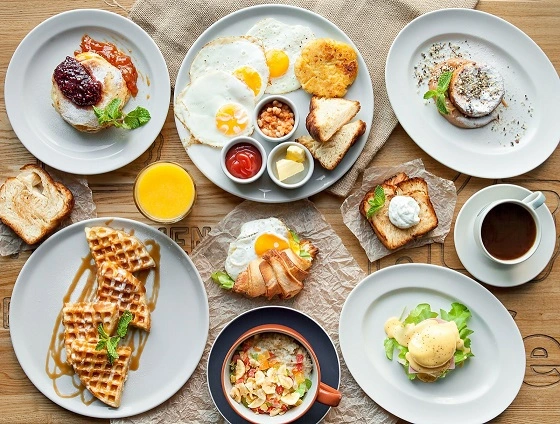Mesa con varios platos de waffles, huevos fritos,croissant, jugo de naranja,pancakes con salsa de frutos rojos y taza de café.